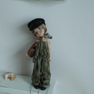 May include: A doll dressed in a black cap, olive green jumpsuit, and black shoes. The doll holds a small, brown book. A small, tan cat figurine rests on a white surface.