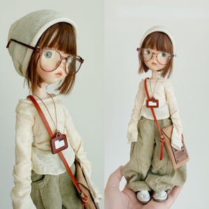 May include: A doll wearing a cream-coloured beanie, round glasses, and a cream-coloured long-sleeved shirt. The doll has short brown hair, olive green trousers, and white trainers. A brown leather bag hangs across the doll's body.