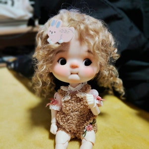 May include: A doll with curly blonde hair wearing a brown fuzzy dress with pink and white floral accents. The doll has a pink bunny hair clip with a white outline.