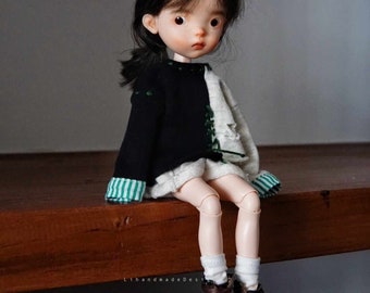 May include: A small doll with black hair, wearing a black and white sweater with green stripes on the sleeves, white socks, and brown boots. The doll is sitting on a wooden surface.