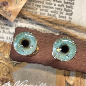 Könnte beinhalten: Zwei tealblaue und schwarze, augenförmige Harzpflaster mit Glitzer und Strasssteinen. Die Pflaster werden auf einem Holzständer präsentiert.