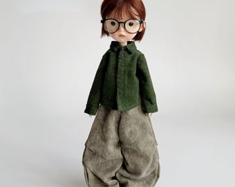 Könnte beinhalten: Eine Puppe mit kurzen, rotbraunen Haaren und einer runden, schwarzen Brille. Die Puppe trägt ein dunkelgrünes Cordhemd und eine übergroße, hellbraune Hose. Die Puppe trägt schwarz-weiße Schuhe.