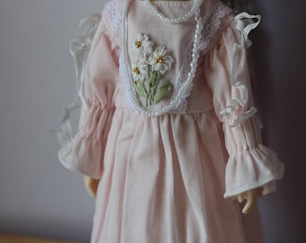 May include: A pink dress with white lace trim and embroidered flowers. The dress has long sleeves with ruffles at the cuffs. A pearl necklace is worn around the neck.