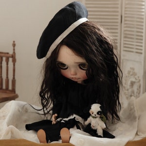Pu&ograve; includere: Una bambola in bianco e nero con lunghi capelli scuri che indossa un berretto nero con un bordo bianco e un vestito nero. La bambola &egrave; seduta su un copriletto bianco e tiene un piccolo peluche bianco e nero.