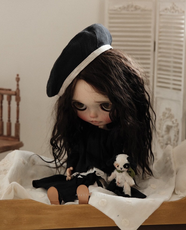 Pu&ograve; includere: Una bambola in bianco e nero con lunghi capelli scuri che indossa un berretto nero con un bordo bianco e un vestito nero. La bambola &egrave; seduta su un copriletto bianco e tiene un piccolo peluche bianco e nero.