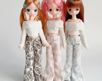 Puede incluir: Tres muñecas de moda con diferentes colores y estilos de cabello. Cada muñeca viste un suéter blanco con hombros descubiertos y pantalones estampados. Los pantalones presentan diseños de estampado animal, floral y pastel. Las muñecas están sobre un fondo blanco.