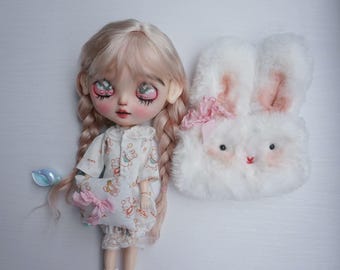 Puede incluir: Una muñeca con largo cabello trenzado rosa claro, vestida con un atuendo blanco con un patrón de osito de peluche. Una bolsa en forma de conejo blanco y esponjoso con acentos rosas está al lado de la muñeca. La muñeca tiene ojos grandes y expresivos y mejillas rosadas.