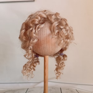 May include: A doll wig with long, curly, light blonde hair. The wig is on a wooden doll head stand.