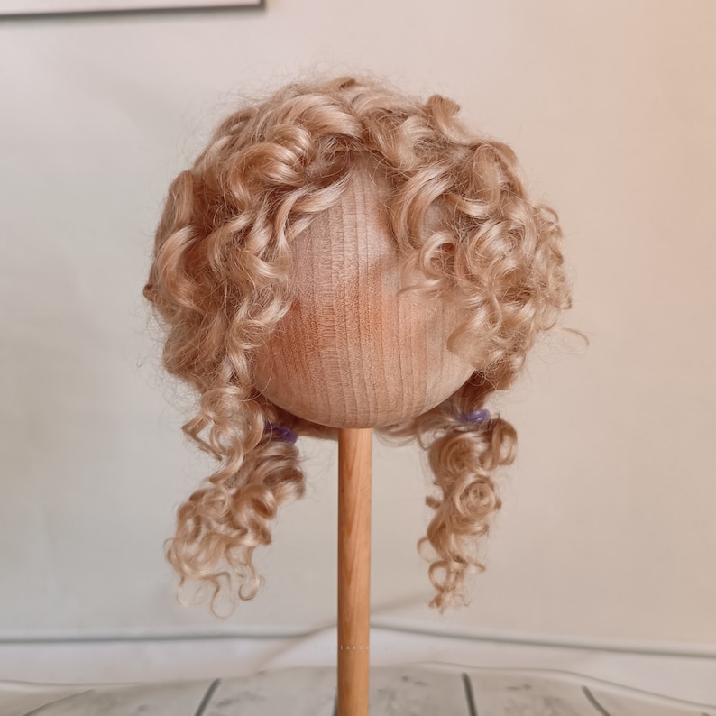 May include: A doll wig with long, curly, light blonde hair. The wig is on a wooden doll head stand.