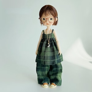 K&ouml;nnte beinhalten: Eine Puppe in einem gr&uuml;n-schwarz karierten Outfit, bestehend aus Oberteil und Hose. Die Puppe hat braunes Haar, eine Brille und eine Halskette. Die Puppe tr&auml;gt braune und wei&szlig;e Schuhe.