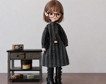 May include: A doll with short brown hair wearing black-rimmed glasses, a dark gray dress with white vertical pinstripes, and black mid-calf boots with buckles. The doll has a small rectangular pendant on a thin cord around its neck.
