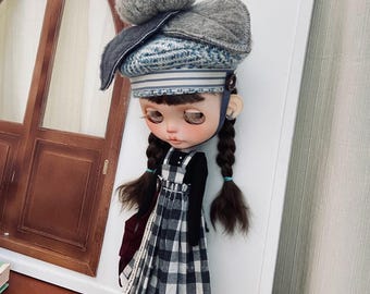 May include: A doll with long brown braids wearing a black long-sleeved shirt, a black and white plaid jumper dress, and a gray and blue knitted hat.