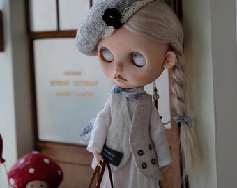 May include: A doll with blonde hair in braids wearing a white dress, a grey vest, and a grey beret. The doll is holding a brown and beige woven bag. The doll is standing in front of a wooden door with a sign that says "Cafe & Cake".