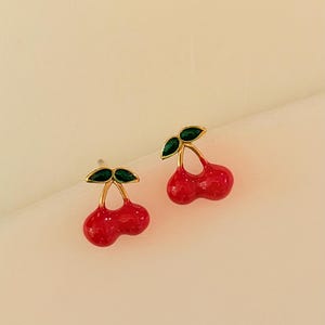 Puede incluir: Un par de pendientes de botón con forma de cereza. Cada pendiente presenta dos cerezas rojas brillantes con un tallo dorado y hojas verdes. Los pendientes están colocados sobre un fondo suave y neutro.