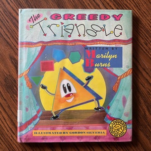 May include: A children's book titled "The Greedy Triangle" with a cartoon triangle character on the cover. The cover features colorful shapes and text, including the author's name, Marilyn Burns, and illustrator, Gordon Silveria.