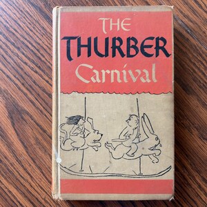 May include: Vintage hardcover book titled "The Thurber Carnival." The cover features a red and beige design with black lettering and illustrations of people riding carousel animals. The book is on a wooden surface.