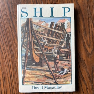 May include: A hardcover book titled "SHIP" by David Macaulay. The cover features an illustration of a ship under construction, with workers and buildings in the background. The book has a white cover with blue lettering.