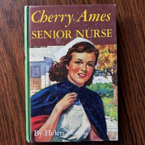 May include: Vintage book cover for "Cherry Ames, Senior Nurse" by Helen Wells. The cover features an illustration of a smiling nurse in a white cap and blue cape. The title is in yellow letters on a maroon background.