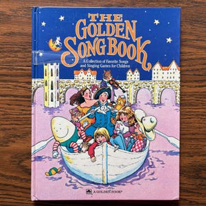 May include: A hardcover book titled The Golden Song Book: A Collection of Favorite Songs and Singing Games for Children. The blue cover features a colorful illustration of various storybook characters rowing a white boat on pink water.