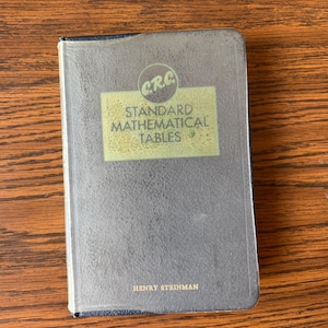 May include: A vintage, navy blue book titled "Standard Mathematical Tables" by Henry Steinman. The book has a clear plastic cover and a gold label with the letters "CRC" in a circle. The book is on a wooden surface.