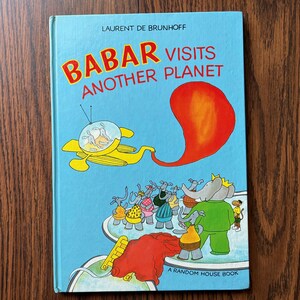 May include: A children's book titled "Babar Visits Another Planet" by Laurent de Brunhoff. The cover features a light blue background with Babar and other elephants in a yellow spaceship and a red, orange, and yellow shape.