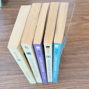 A collection of five paperback books by Lloyd Alexander are lined up on a wooden surface. The spines of the books display titles such as The High King, Taran Wanderer, The Black Cauldron, and two others.