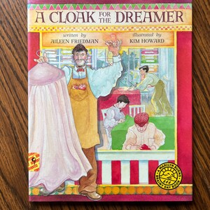 May include: A children's book cover titled "A Cloak for the Dreamer" with illustrations. The cover features a tailor holding a pink cloak, with other figures in the background. The author is Aileen Friedman, and the illustrator is Kim Howard.