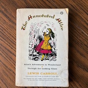 May include: A vintage paperback book titled "The Annotated Alice" with a cover illustration of Alice in Wonderland. The cover features a drawing of Alice surrounded by playing cards. The book is by Lewis Carroll.
