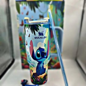 Peut inclure: Un gobelet Stanley bleu avec un motif de personnage de dessin animé. Le gobelet a une paille bleue et un couvercle. Le motif comprend un personnage bleu avec de grandes oreilles, sur fond de papillons et de feuillage. Le logo Stanley est visible.