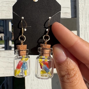 May include: A pair of novelty earrings featuring miniature glass bottles filled with colorful candies. Each bottle has a cork stopper and is suspended from a gold-colored hook. The candies are red, blue, and yellow.