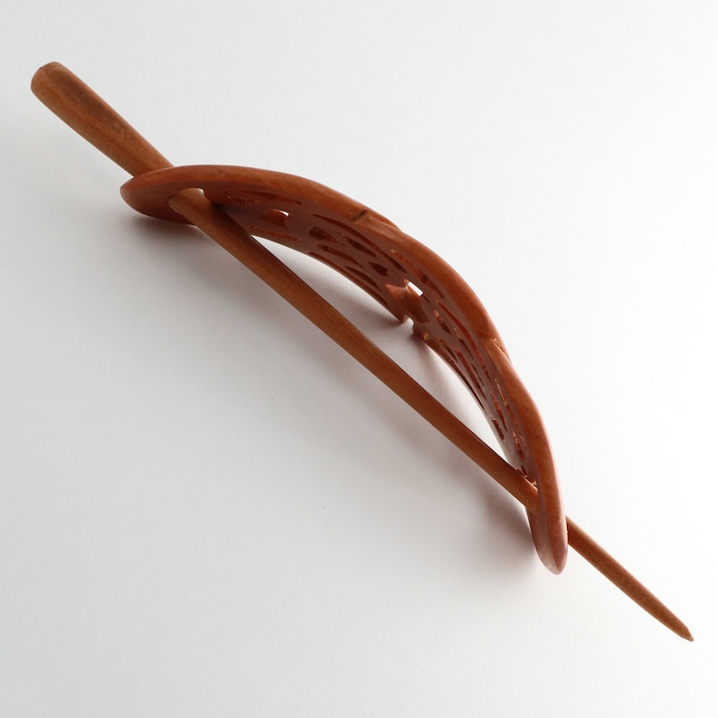 May include: A wooden hair stick with a carved, curved design. The hair accessory is a warm brown color and features a long, pointed pin. The decorative element has an openwork pattern, adding visual interest to the hair pin.