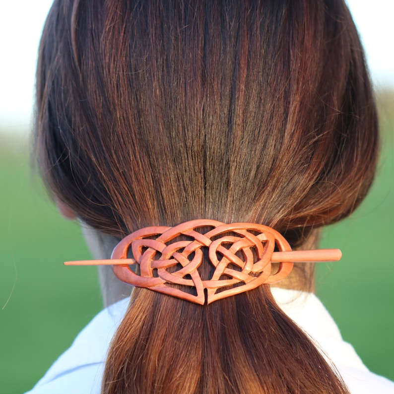 May include: A wooden hair barrette with a Celtic knot heart design. The barrette is a warm reddish-brown color and is inserted into a ponytail. The hair accessory is a unique and stylish way to secure hair.