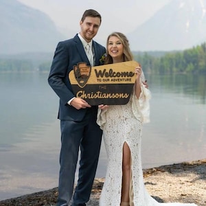 May include: A couple poses for a wedding photo holding a wooden sign that says "Welcome to our adventure, The Christiansons". The sign has a wood-burned image of a national park logo. The couple is standing in front of a lake with mountains in the background.
