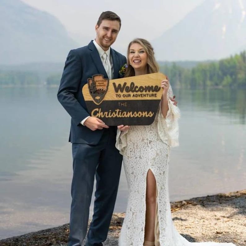 May include: A couple poses for a wedding photo holding a wooden sign that says "Welcome to our adventure, The Christiansons". The sign has a wood-burned image of a national park logo. The couple is standing in front of a lake with mountains in the background.