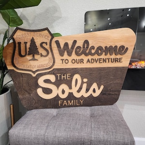 May include: A wooden sign with a brown and light wood finish. The sign is shaped like a rounded rectangle with a beveled edge. The sign has a carved US Forest Service logo on the left side and the text "Welcome to our adventure" on the right side. Below the text is the name "The Solis Family" in a decorative font.
