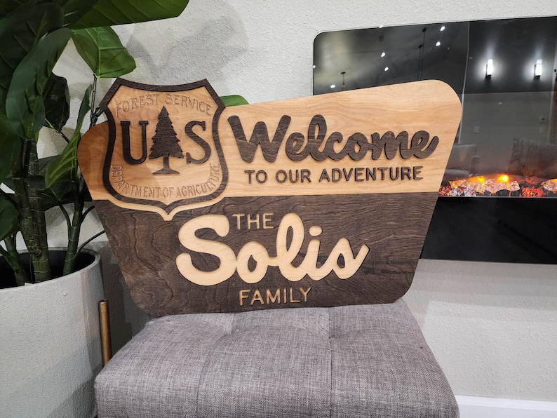 May include: A wooden sign with a brown and light wood finish. The sign is shaped like a rounded rectangle with a beveled edge. The sign has a carved US Forest Service logo on the left side and the text "Welcome to our adventure" on the right side. Below the text is the name "The Solis Family" in a decorative font.