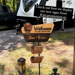 May include: A white camper with the word "Imagine" on the side, parked in a grassy area. There are wooden signs with the text "Welcome to our adventure" and "Jen + Eric".