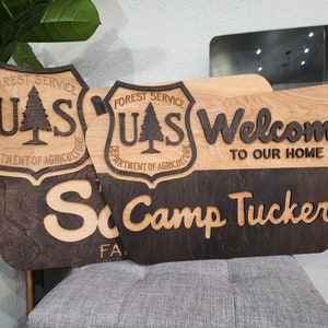 May include: Two wooden signs with a dark brown stain and a light wood stain. The signs feature the US Forest Service logo, the text "Welcome to our home", and the text "Camp Tucker".