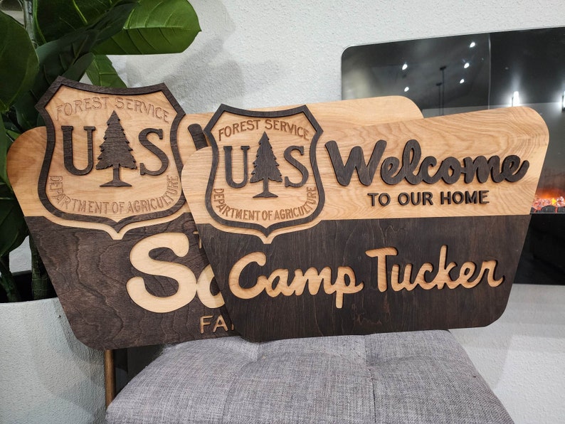 May include: Two wooden signs with a dark brown stain and a light wood stain. The signs feature the US Forest Service logo, the text "Welcome to our home", and the text "Camp Tucker".