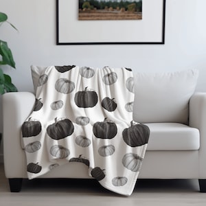 May include: A white throw blanket with a black and white pumpkin pattern. The blanket is folded and draped over a white couch.