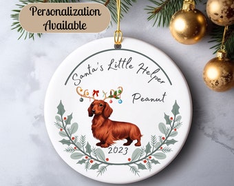 May include: A white ceramic Christmas ornament featuring a dachshund dog wearing a reindeer antler headband with red and green ornaments. The ornament has the text "Santa's Little Helper Peanut 2023" on it.