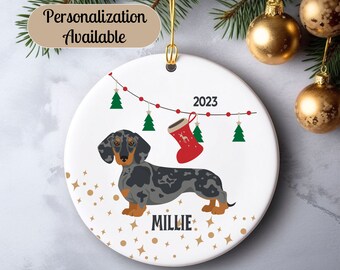 May include: A white ceramic Christmas ornament featuring a black and blue Dachshund dog with the name "Millie" and the year "2023". The ornament is decorated with a string of red and white Christmas lights, a red stocking, and gold glitter.