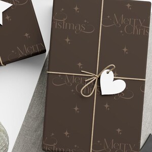 Neutral Christmas gift wrapped in deep mocha brown paper with minimalist “Merry Christmas” text and stars, tied with simple beige twine, nestled in pine branches
