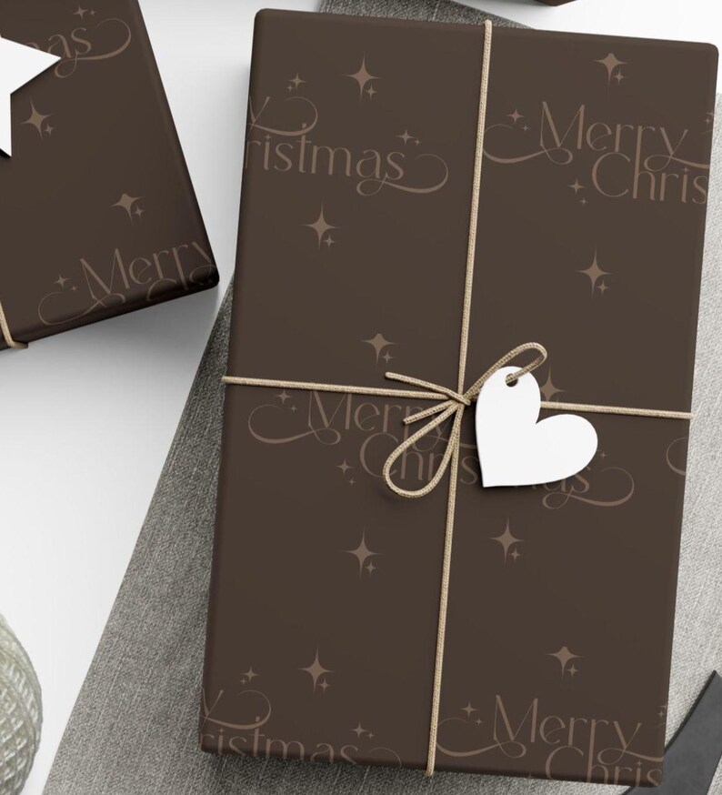 Neutral Christmas gift wrapped in deep mocha brown paper with minimalist “Merry Christmas” text and stars, tied with simple beige twine, nestled in pine branches