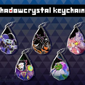 Könnte beinhalten: Fünf tropfenförmige Acryl-Schlüsselanhänger mit farbenfrohen Charakterillustrationen. Jeder Schlüsselanhänger hat eine silberne Kugelkette. Der Text "shadowcrystal keychains" ist oben im Bild in einer Pixel-Schriftart zu sehen.