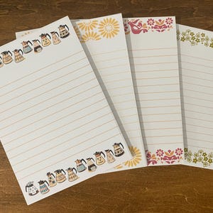 May include: Four sheets of white lined stationery paper. The first sheet features a border of colorful coffee pots. The second sheet has yellow floral patterns. The third sheet displays pink and red folk-style motifs. The fourth sheet has green floral borders.