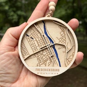 May include: A circular wooden ornament featuring a laser-cut map of Fredericksburg with a blue inlay representing a river. The piece includes the text FREDERICKSBURG and the coordinates 38 18 6.5 N, 77 28 15 W, finished with a jute twine and wooden beads.