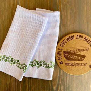 May include: Two white waffle-weave kitchen towels with a green floral border are next to a round cork coaster. The coaster has the text "BOWLS, BAKEWARE AND BRAGGING RIGHTS" around a graphic of three bowls.