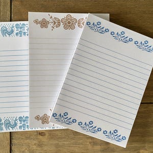 May include: Three rectangular sheets of white stationery paper with horizontal blue lines. The left sheet features blue rooster and floral motifs. The center sheet has brown floral borders. The right sheet displays blue floral patterns at the top and bottom.