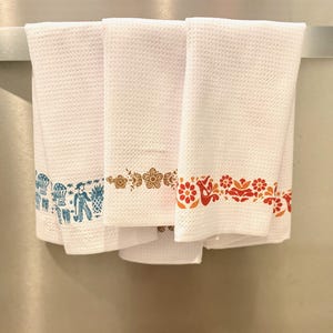 May include: Three white waffle-weave kitchen towels hanging on a silver bar. Each towel has a decorative border in different colors: blue, brown, and red. The blue border features a farmer and other illustrations. The towels are clean and neatly folded.